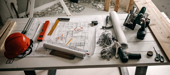 Construction tools, safety gear, and blueprints arranged on a table, featuring a hard hat, drill, measuring tape, hammer, safety gloves, and various other tools, set in a partially finished workspace.